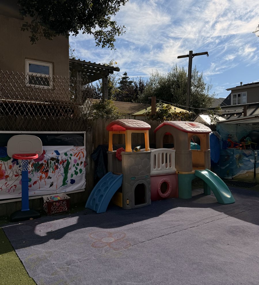 The Pickles play yard on a sunny afternoon — a climbing structure with two slides, a toddler basketball hoop, and a long banner the children painted, under an open blue sky.